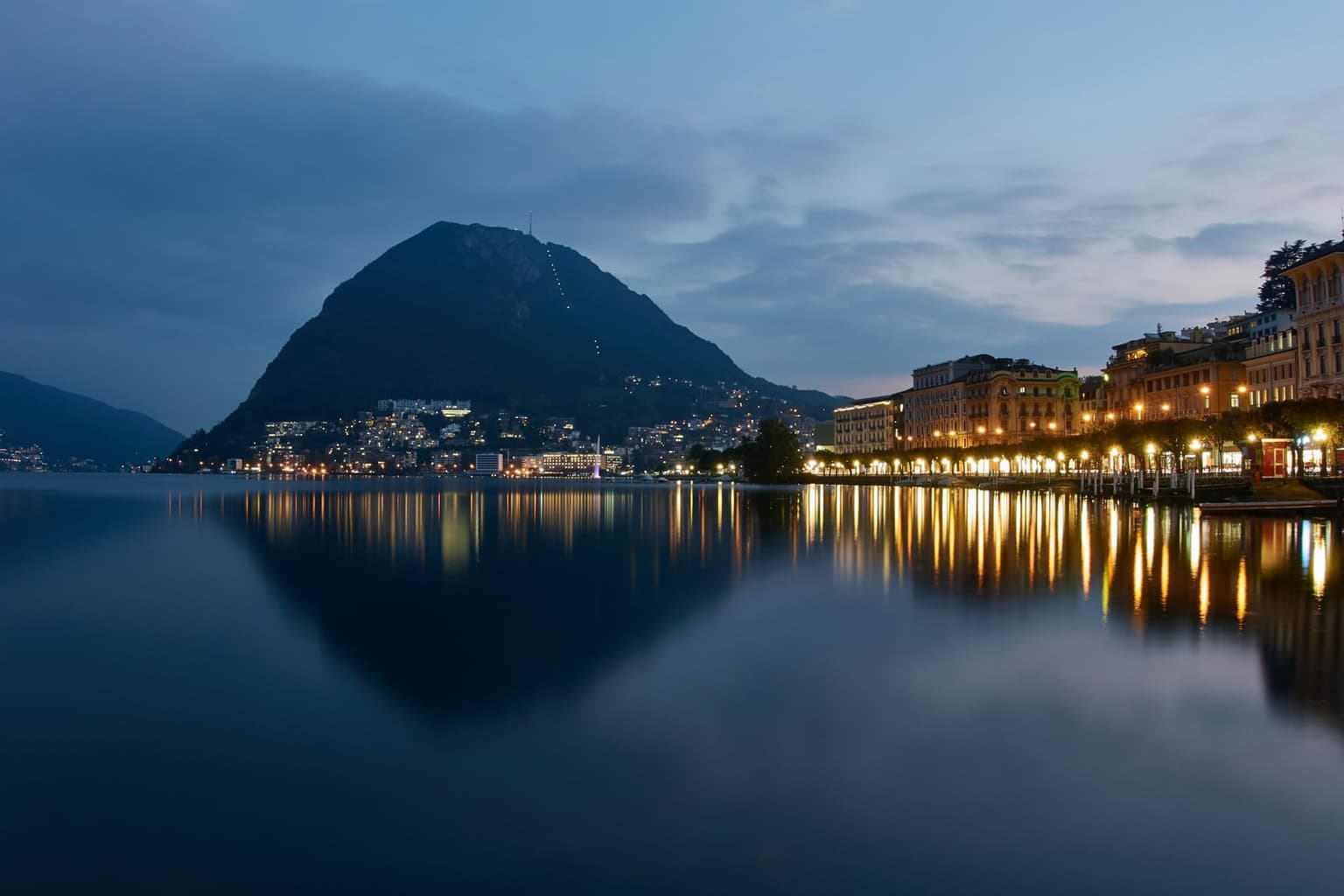 Lugano's lakefront at dusk, lit hotels along the promenade on the right and the steep cone of Monte San Salvatore rising across the still lake