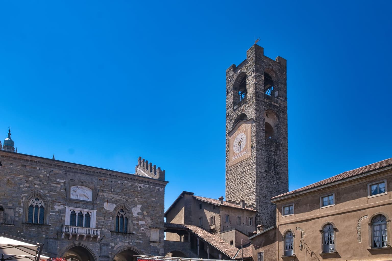 Piazza Vecchia in Bergamo's upper town, the stone civic tower with its big clock face rising behind old palazzi, and the Palazzo della Ragione on the left with Gothic windows and a carved lion