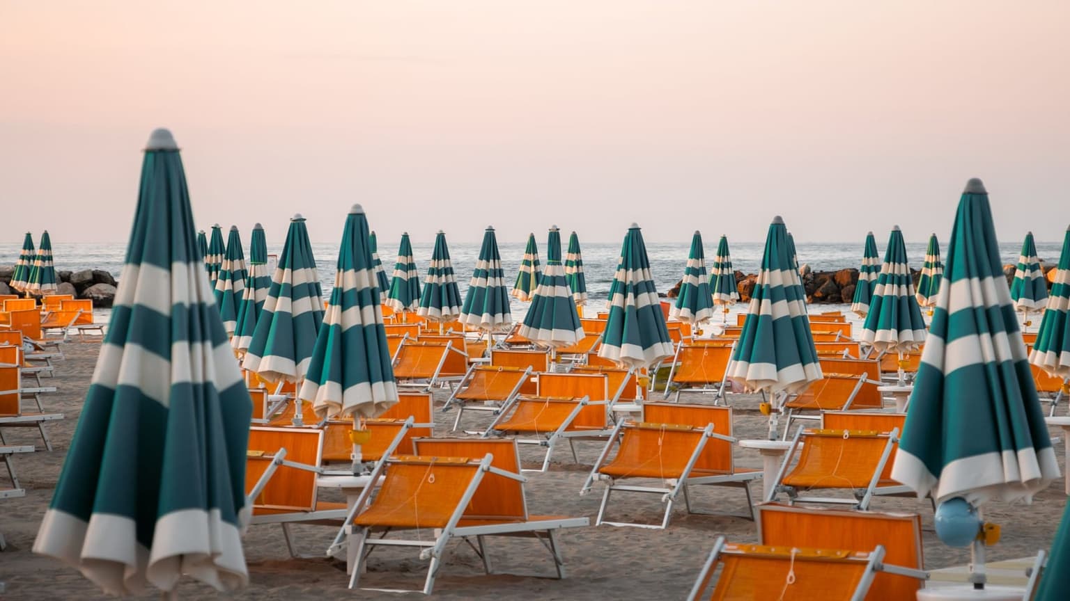 Rows of orange sun loungers and closed teal-and-white striped umbrellas lined up on a sandy beach at sunset, running out toward the sea and horizon