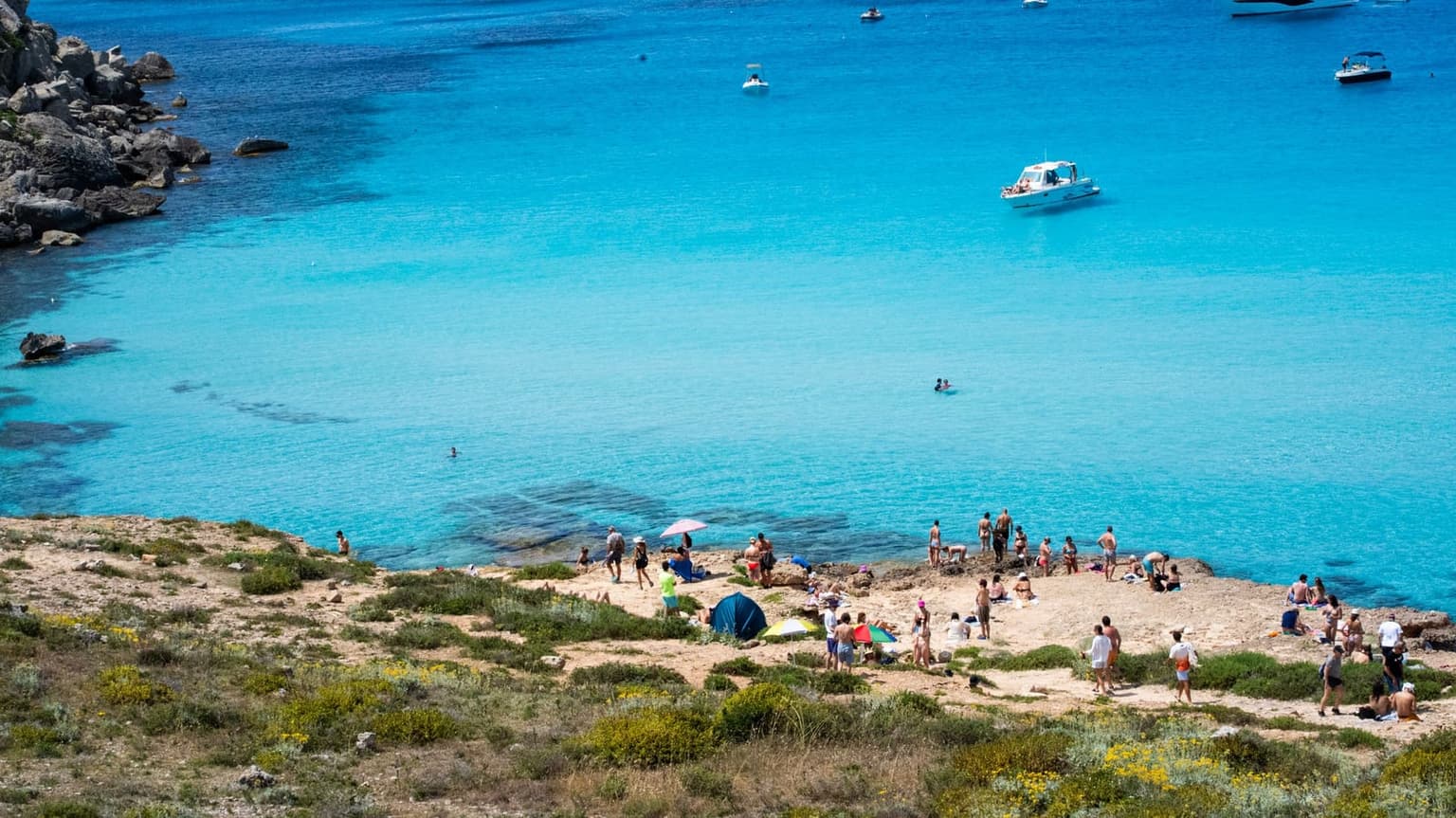 A turquoise cove at Calamosche seen from a rocky, scrub-covered headland, with a motorboat and swimmers in the water and sunbathers with a couple of umbrellas on the sand below