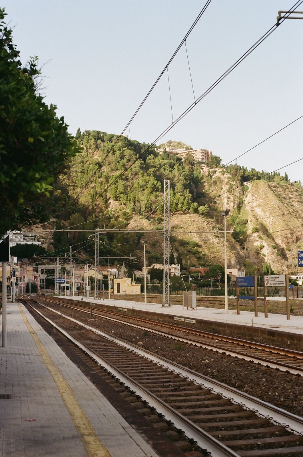 An empty regional train platform in Sicily with tracks curving away beneath overhead power lines, a green hillside and a hilltop hotel building in the background