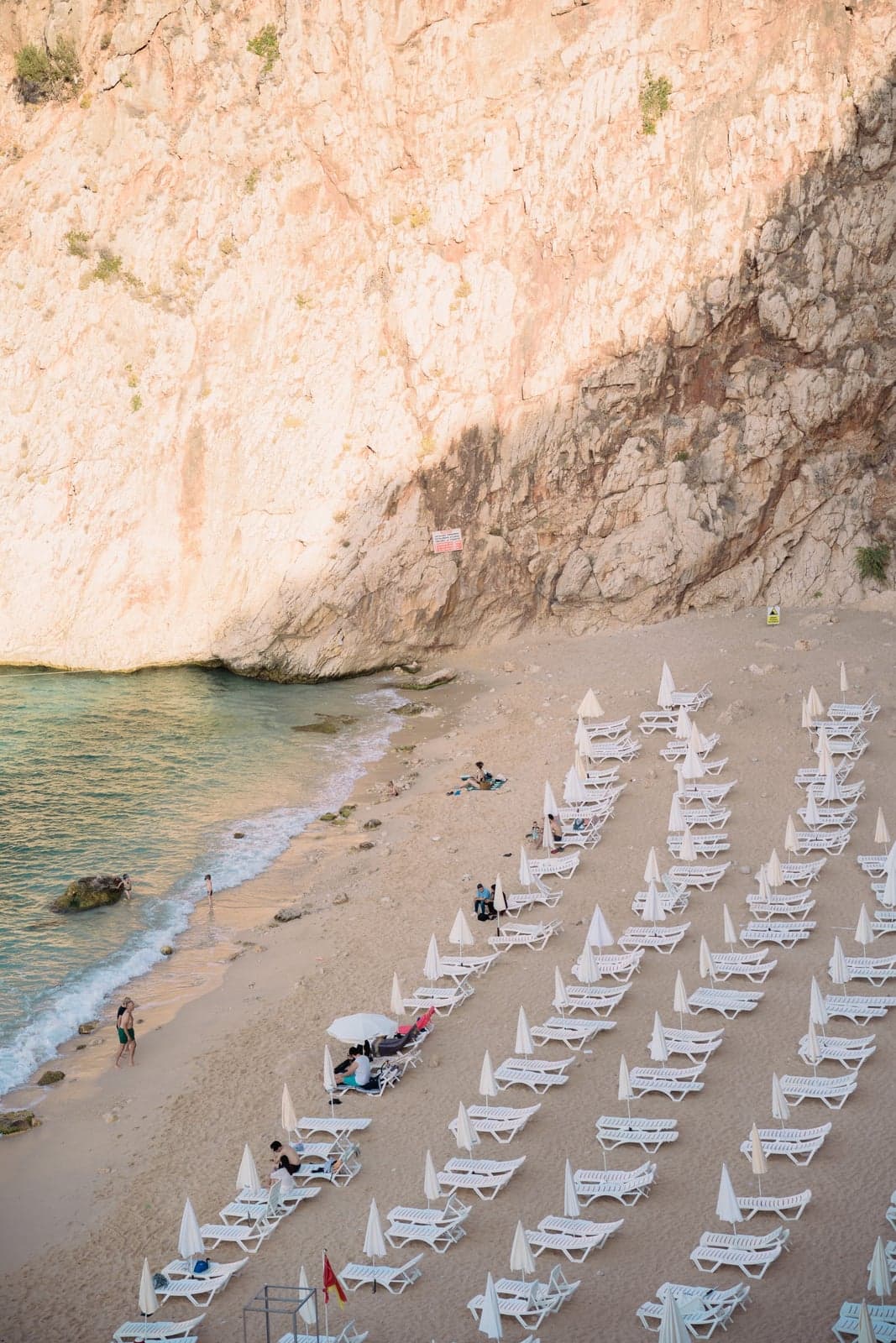 An aerial view of a cove beach: white sun loungers and umbrellas fill one stretch of sand against the cliff base while the rest of the shoreline and turquoise water stay open and empty