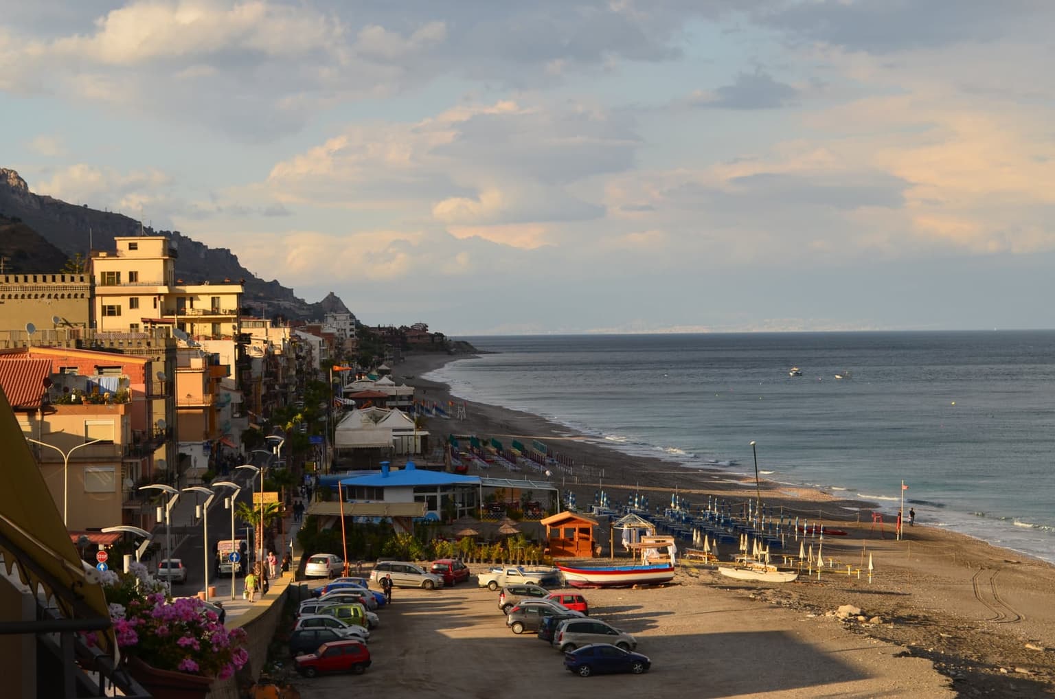 Letojanni's beachfront seen from above the town at evening: a fishing boat pulled up on the sand, rows of beach umbrellas farther along the shore, and the coastline curving toward a distant headland under warm light