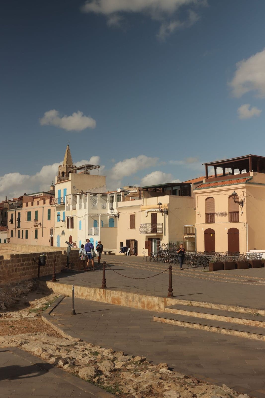 Marzamemi's small seaside piazza lined with low pastel Sicilian buildings and outdoor café tables, a bell tower with a cross rising above the rooftops