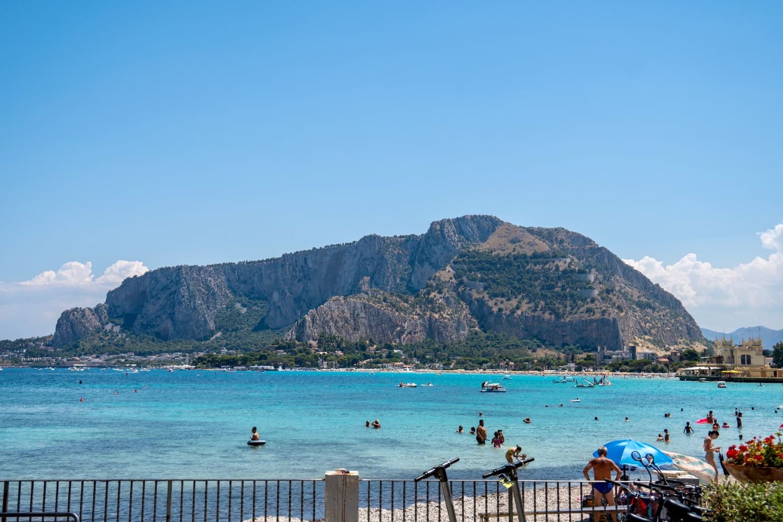 Mondello's curved bay with Monte Pellegrino rising behind it, swimmers in the turquoise water and beach umbrellas lining the far shore