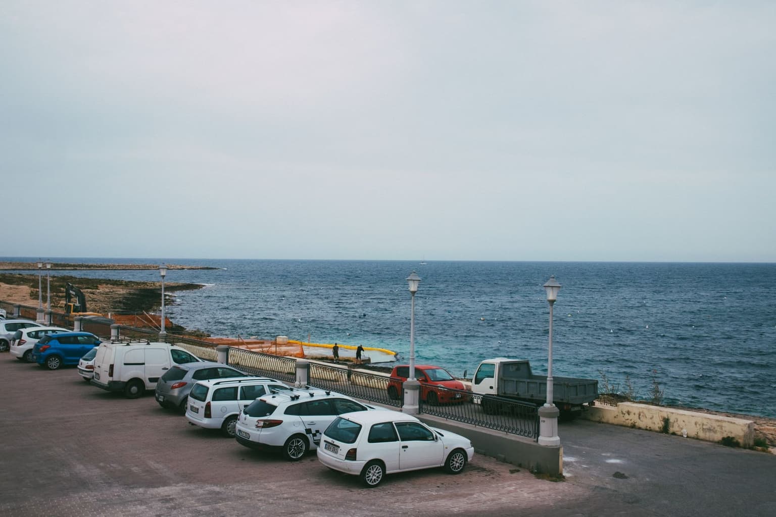 Cars parked in a row along a paved seaside promenade with ornamental iron railings and lamp posts, the open sea beyond, no painted parking lines or ticket machine visible