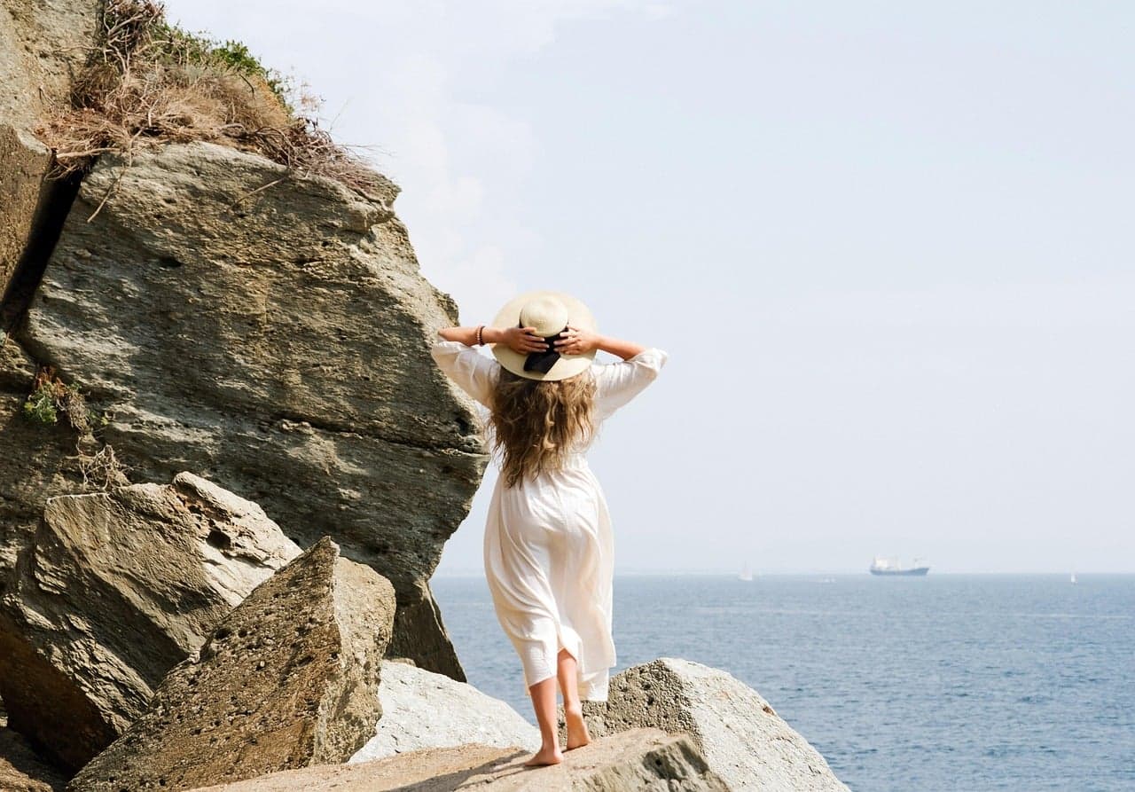 A woman stands at the edge of pale, chalky rock formations at Punta Bianca overlooking the sea, a cargo ship visible on the horizon, no built structures in sight