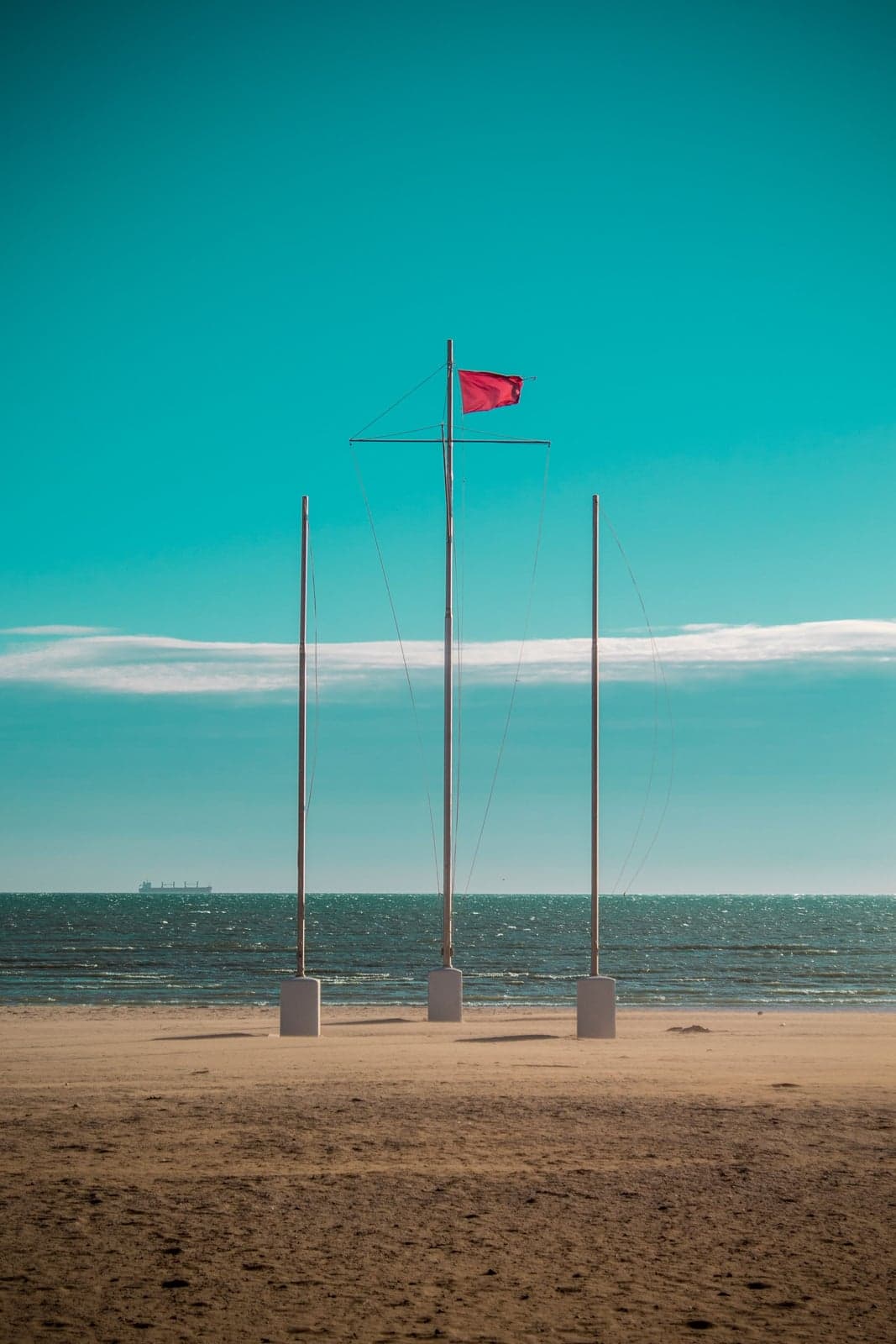 A red warning flag flying from a pole on an empty sandy beach, with a calm teal-blue sea and a cargo ship visible on the horizon