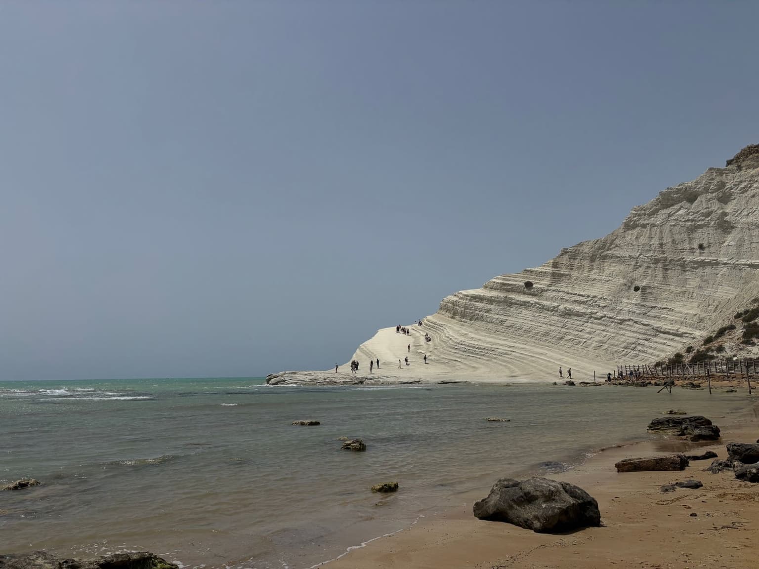 Scala dei Turchi's white limestone stair-terraces seen from beach level, with people climbing and standing on the steps for scale, the sea in the foreground