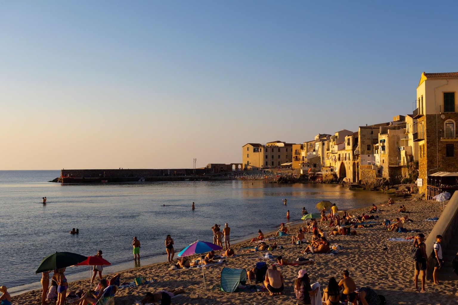 Cefalù's old town at golden hour, pastel houses rising directly from a sandy beach crowded with sunbathers and colorful beach umbrellas, calm sea in the foreground