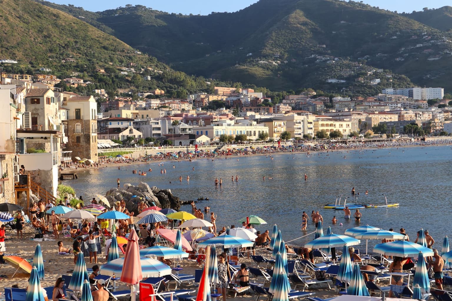 An elevated view of a packed Sicilian beach at Cefalù in summer, dense rows of umbrellas and sunbeds lining the sand, crowds in the shallow water, the town's hillside behind.