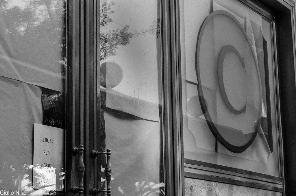 A handwritten "chiuso per ferie" (closed for the holidays) sign taped inside a shop's glass door, reflections of trees visible in the glass.