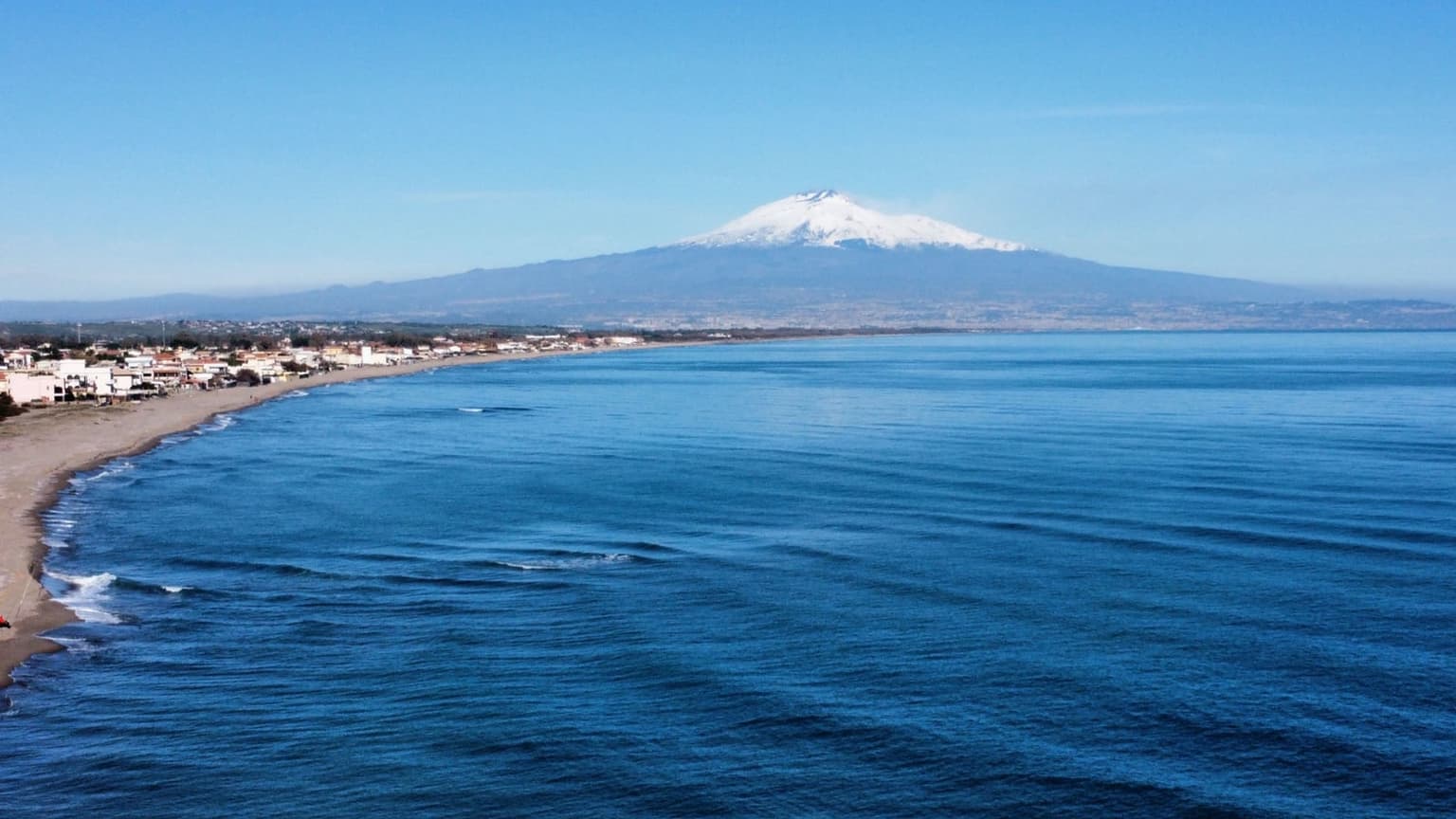 A snow-capped Mount Etna rises above a sunlit coastal town and calm blue sea, seen from an elevated angle looking south along the shoreline.
