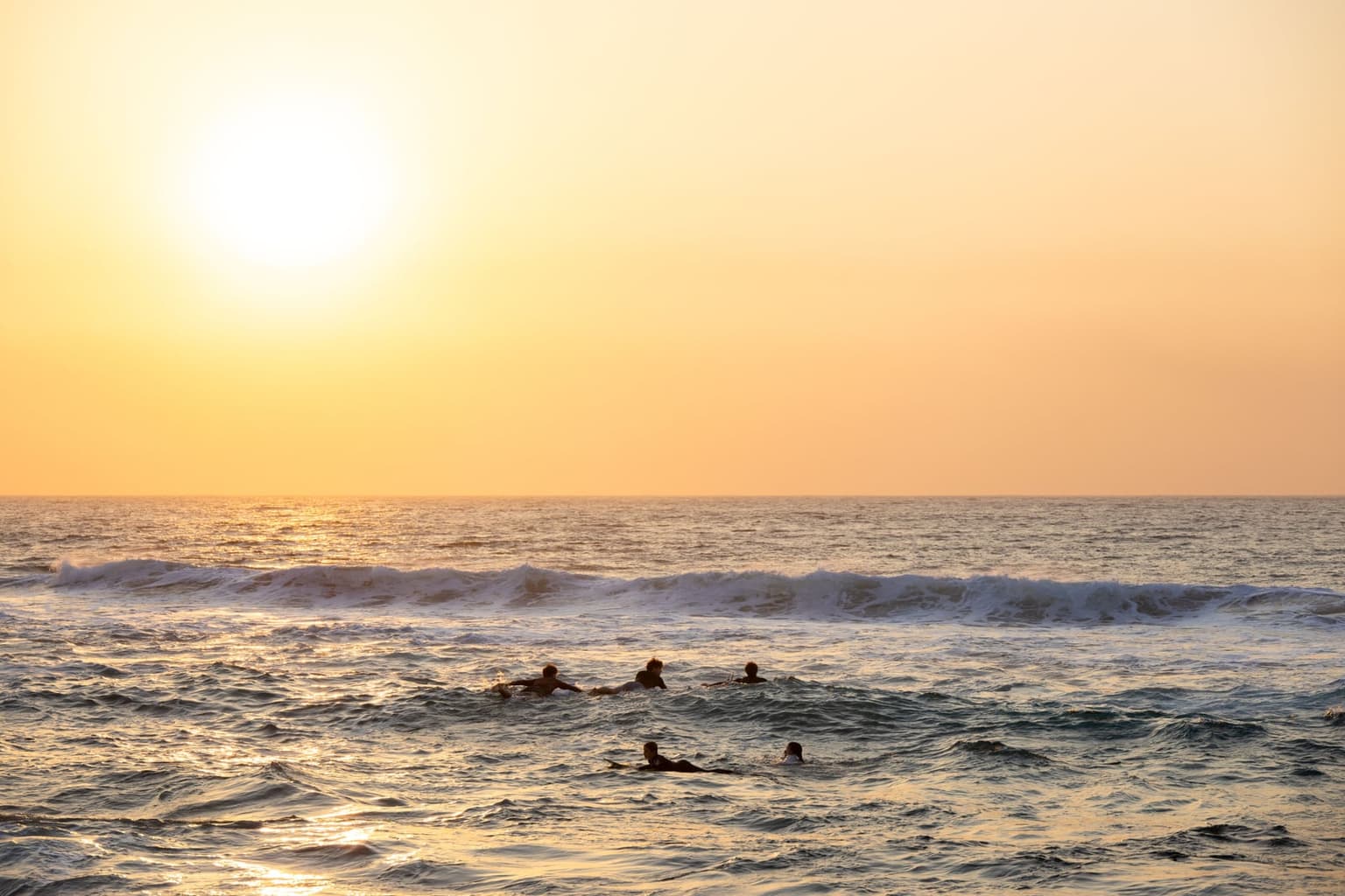 Several swimmers float in the sea past breaking waves under a hazy golden sun low on the horizon.