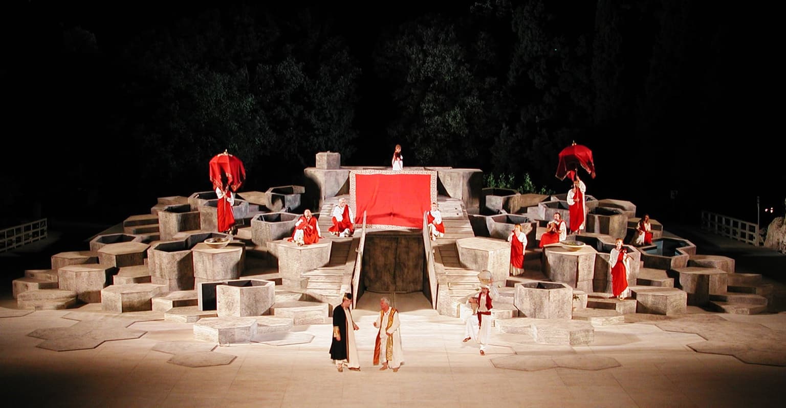Actors in red-and-white robes perform on a purpose-built stage set that fills the ancient stone seating bowl of Siracusa's Teatro Greco at night, floodlit for a classical drama production.