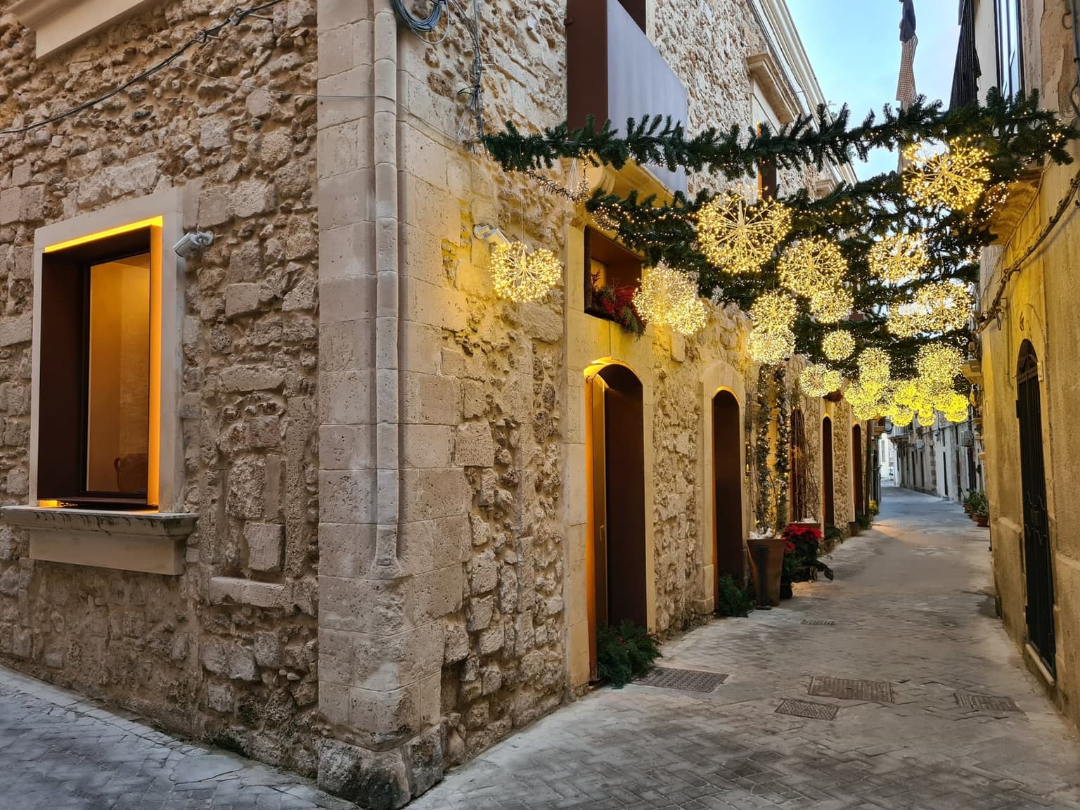 A narrow Siracusa alley strung with lit Christmas garlands between the balconies at dusk, empty and quiet, stone walls glowing warm against the fading blue sky.