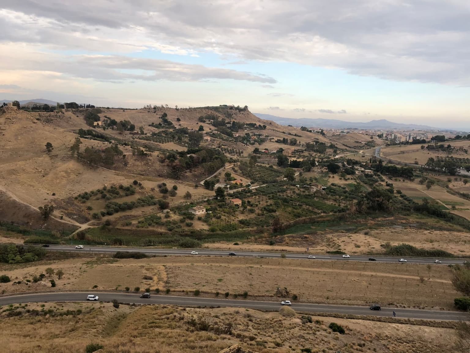 Two roads with light traffic run below dry hills of olive groves and scattered farmhouses near Agrigento, seen from the church of San Nicola, with the city's apartment blocks on the right.