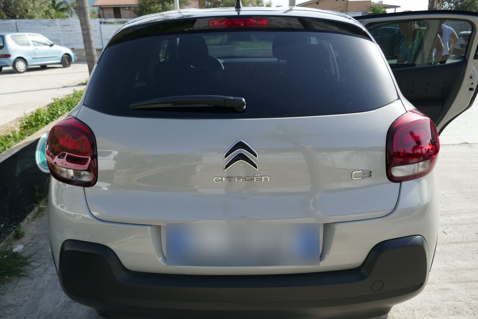 A light gray Citroën C3 seen from behind in a paved lot, its rear door open and the license plate blurred.
