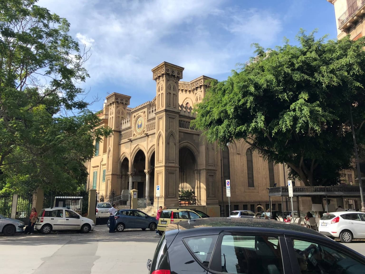 Small cars parked nose to tail along a curb in Palermo, in front of a sandstone neo-Gothic building with pointed arches and tall corner towers, as a dark hatchback waits in the lane in the foreground with its driver at the wheel.
