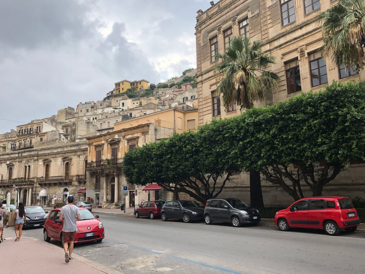 Small cars, two of them Fiat Pandas, parked nose to tail under trimmed trees along a street in Modica, with baroque facades beside them and the old town climbing the hill behind.