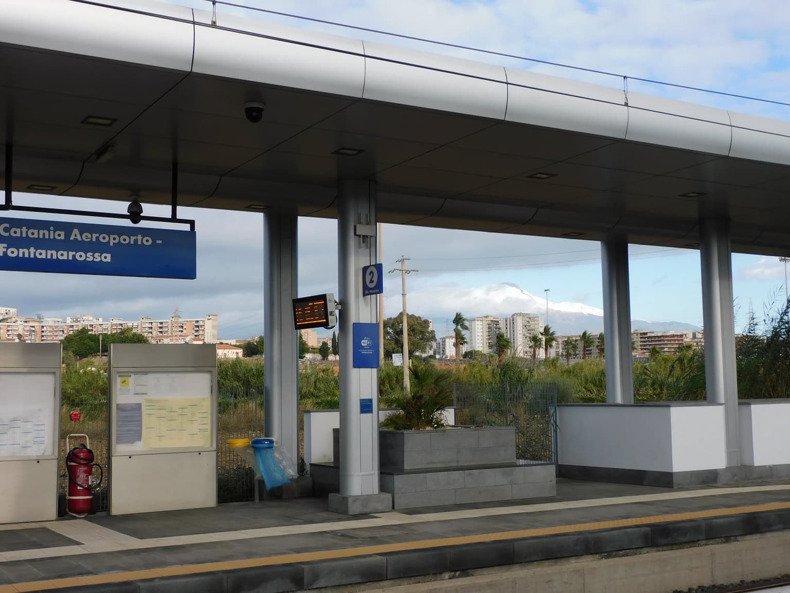 The Catania Aeroporto-Fontanarossa train platform, its blue overhead sign naming the station, with snow-capped Mount Etna rising above apartment blocks and palm trees on the skyline beyond the tracks.