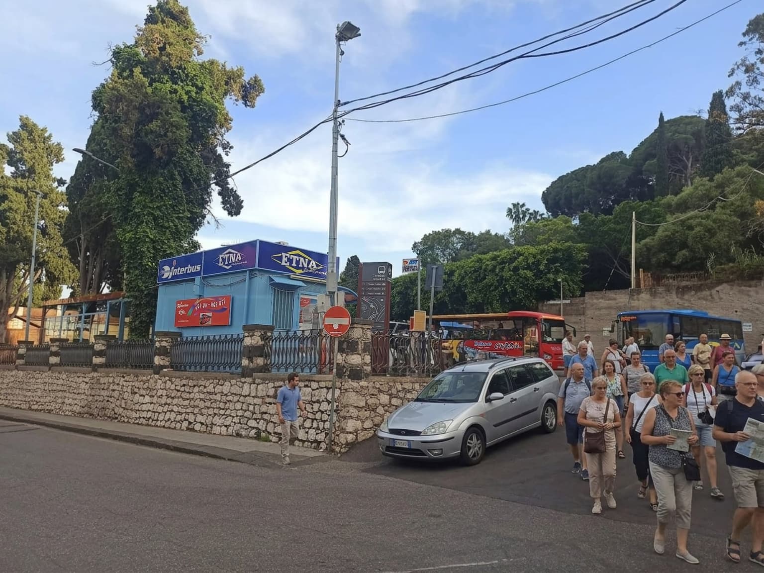 Street-level view of the blue Interbus / Etna Trasporti bus terminal building in Taormina, with a red Hop On Hop Off tour sign on its facade, a dark signpost reading Terminal Interbus with arrows toward Teatro Antico, Porta Messina, and Porta Catania, buses parked along the curb, and pedestrians crossing the road past a low stone wall in the foreground.