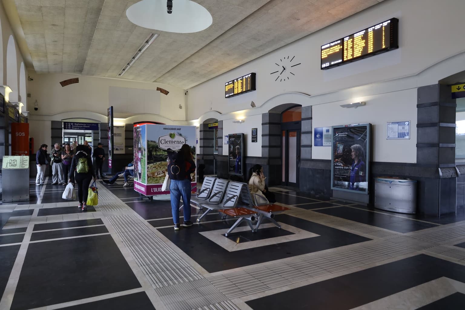 The interior of Catania Centrale station with an illuminated departures board on the wall, travelers with luggage in the foreground.