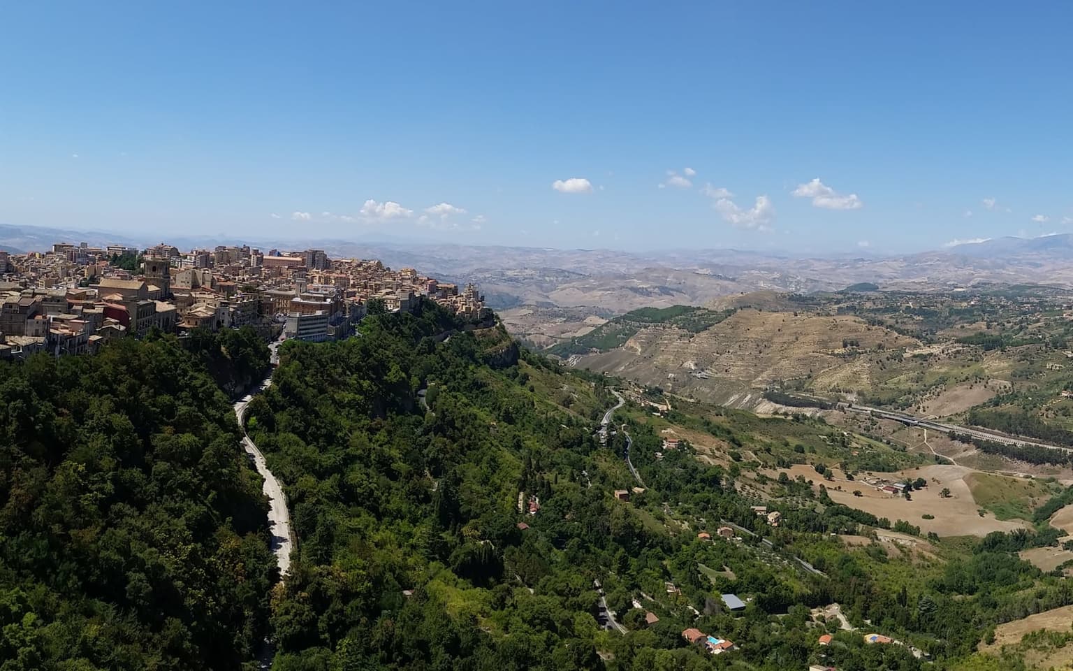 An elevated view over the hill town of Enna and the valley below, with a paved switchback road visibly cutting down the green hillside into the valley, dry rolling hills and a distant highway viaduct in the background under a clear blue sky.