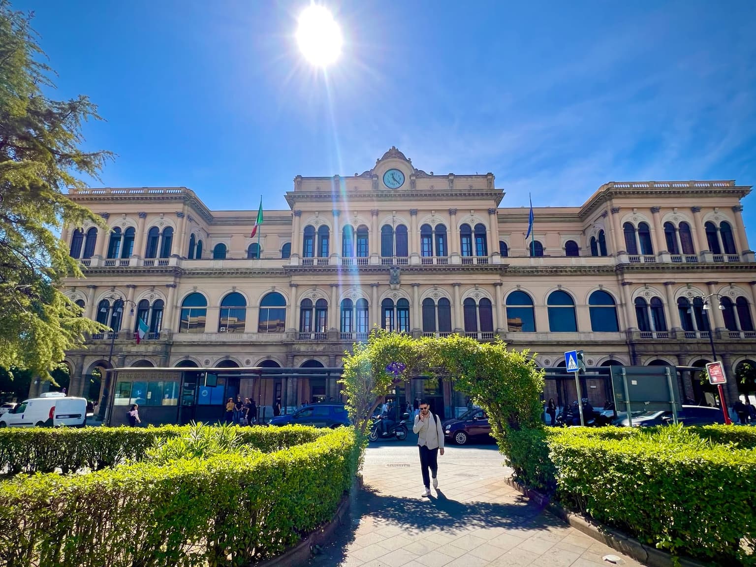 A sunlit, low-angle view of the Palermo Centrale station facade, a symmetrical sandstone building with a central pedimented clock tower and rows of arched windows, a pedestrian walking through a trimmed hedge archway in the foreground.