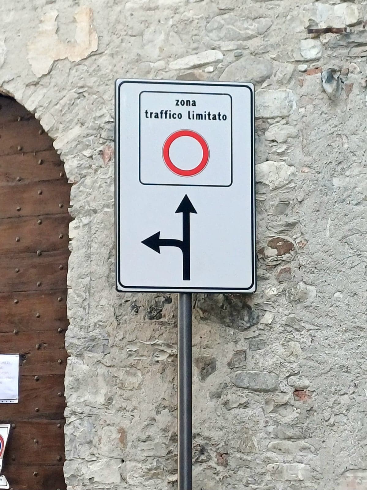 A standard Italian "zona traffico limitato" (limited traffic zone) road sign mounted on a pole against a rough stone wall, showing the red-ringed restricted-access symbol above straight-ahead and left-turn arrows.