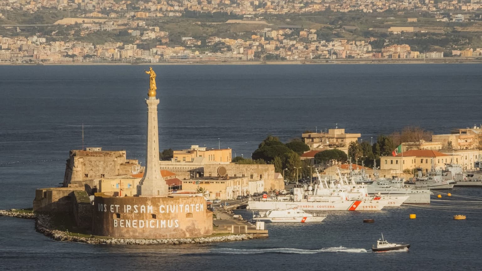 The Madonna della Lettera monument on its column at the mouth of Messina's harbor, Coast Guard vessels docked below and the city spread along the waterfront