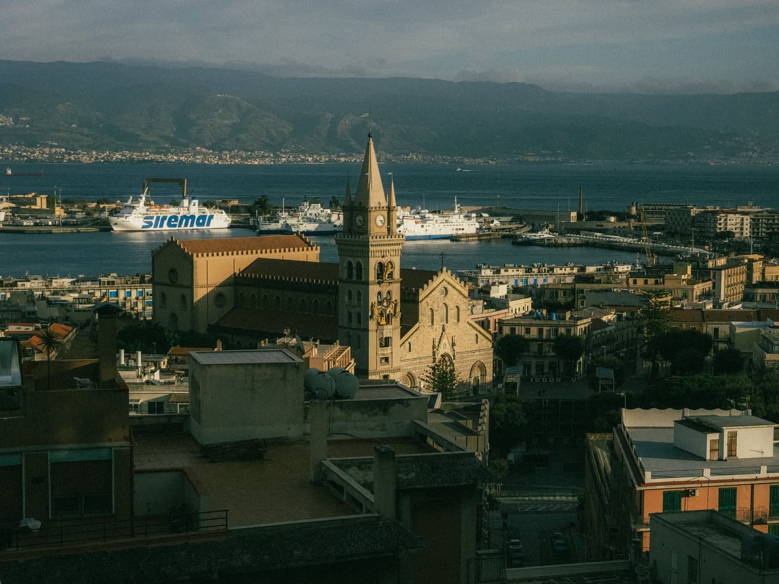 Aerial view of Messina's old town, the Duomo bell tower rising above the rooftops with the harbor and a docked ferry below, Calabria's coastline visible across the Strait of Messina
