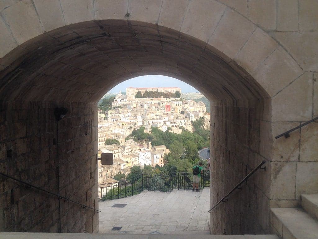 A stone archway framing the descent into Ragusa Ibla, steps leading through the gate to a railed overlook with the town's rooftops spread out below.