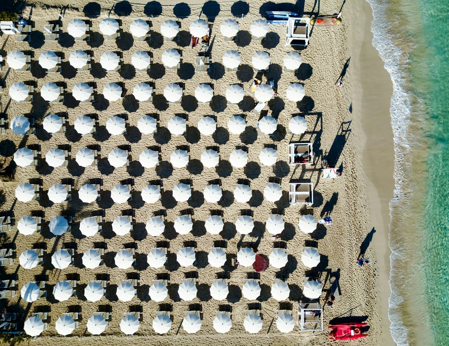 Aerial view straight down onto rows of white beach umbrellas and lounge chairs on tan sand, arranged in graded, numbered rows running back from the turquoise shoreline.