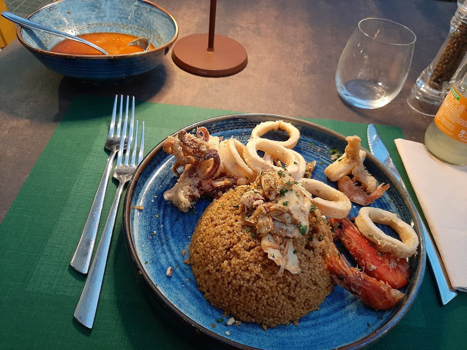 A plate of San Vito fish couscous: a mound of coarse yellow semolina topped with fish and shellfish, fried calamari and shrimp alongside, with a bowl of fish broth on the table.