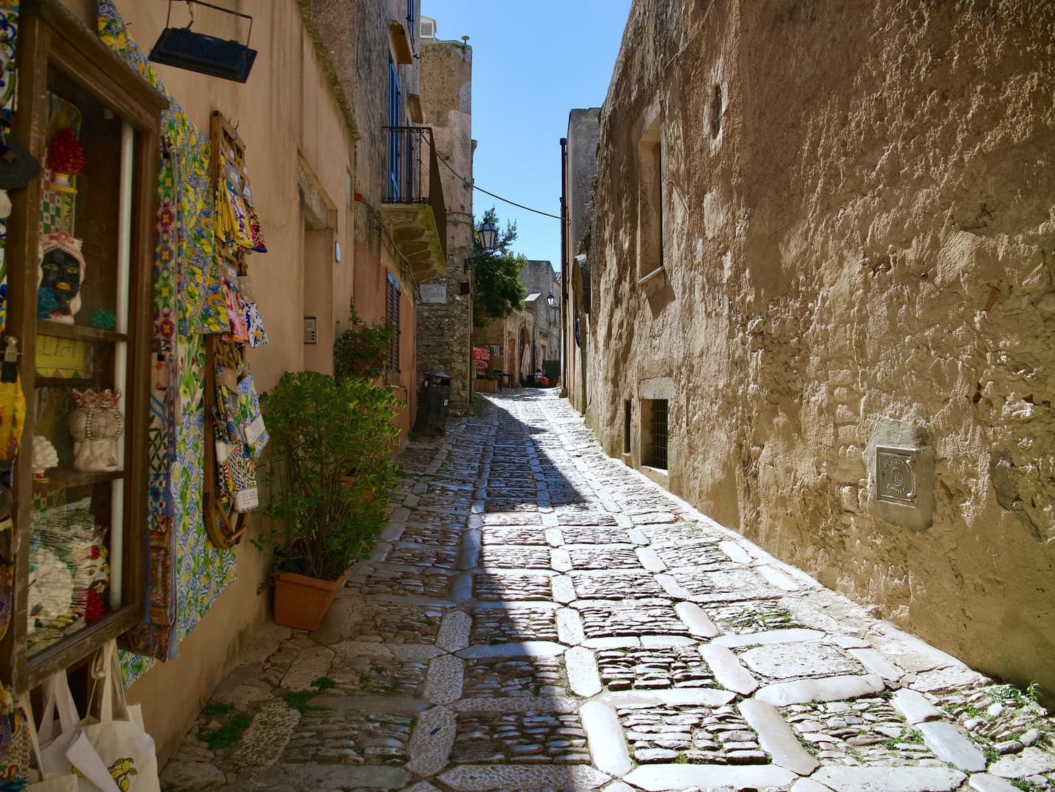 A narrow, sunlit stone-paved lane between old sandstone buildings in Erice, with colorful ceramics and textiles hanging in a shop window and the lane sloping into shadow ahead.