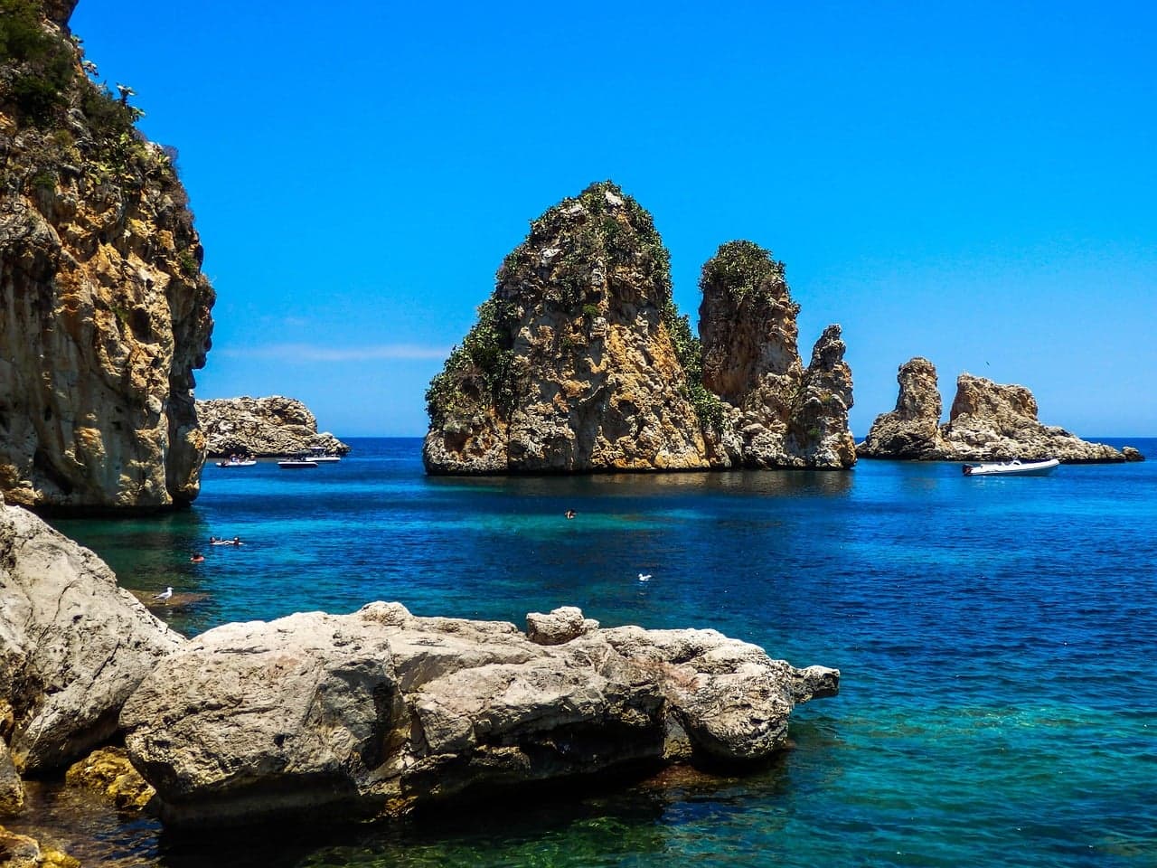 Two dark, weathered rock towers rising straight out of clear turquoise water at sea level, with a smaller rock formation beyond and small boats and swimmers near their base.
