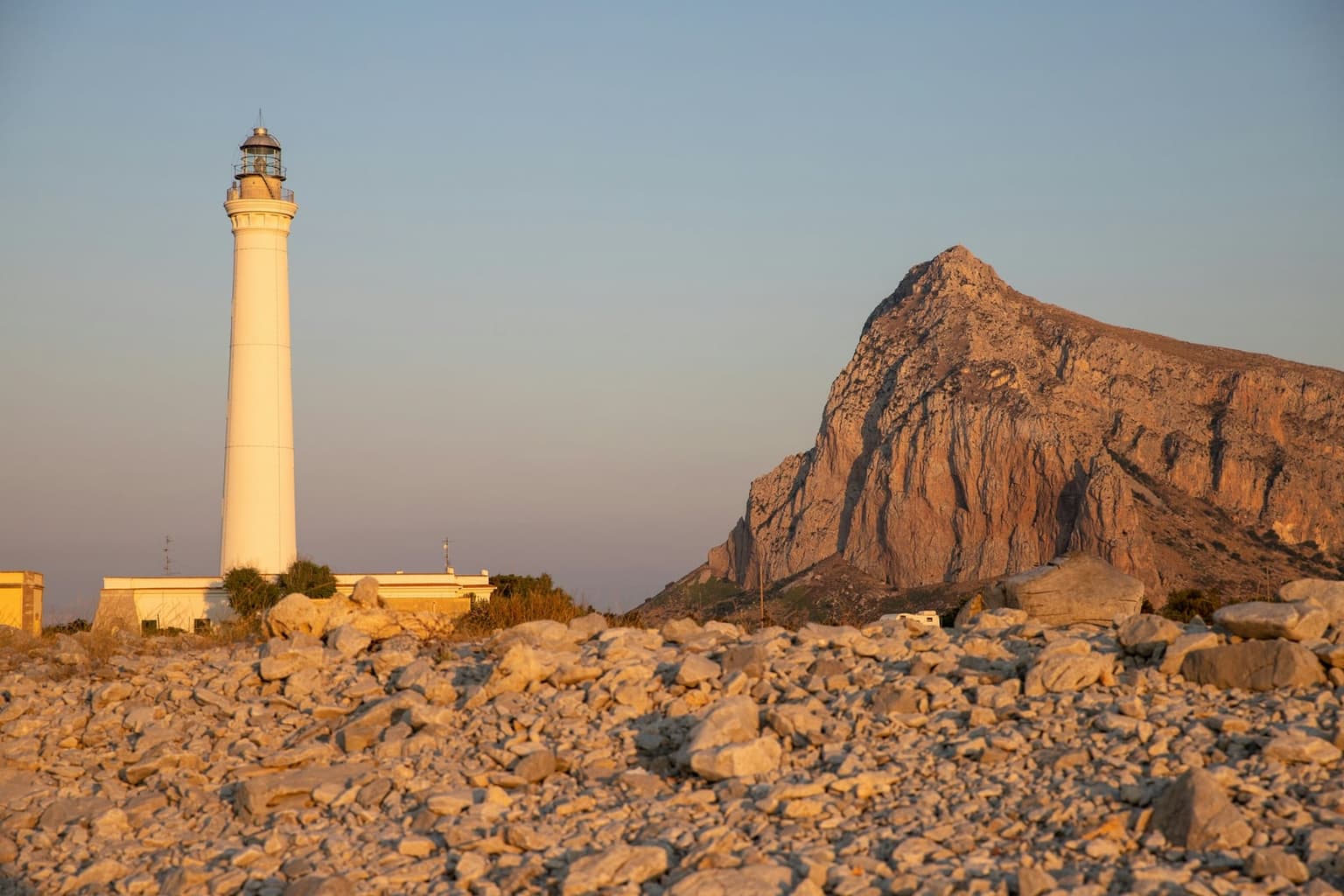 A tall white lighthouse tower lit by low sun beside a squat stone outbuilding, with a boulder-strewn rocky foreground and a mountain peak rising behind it.