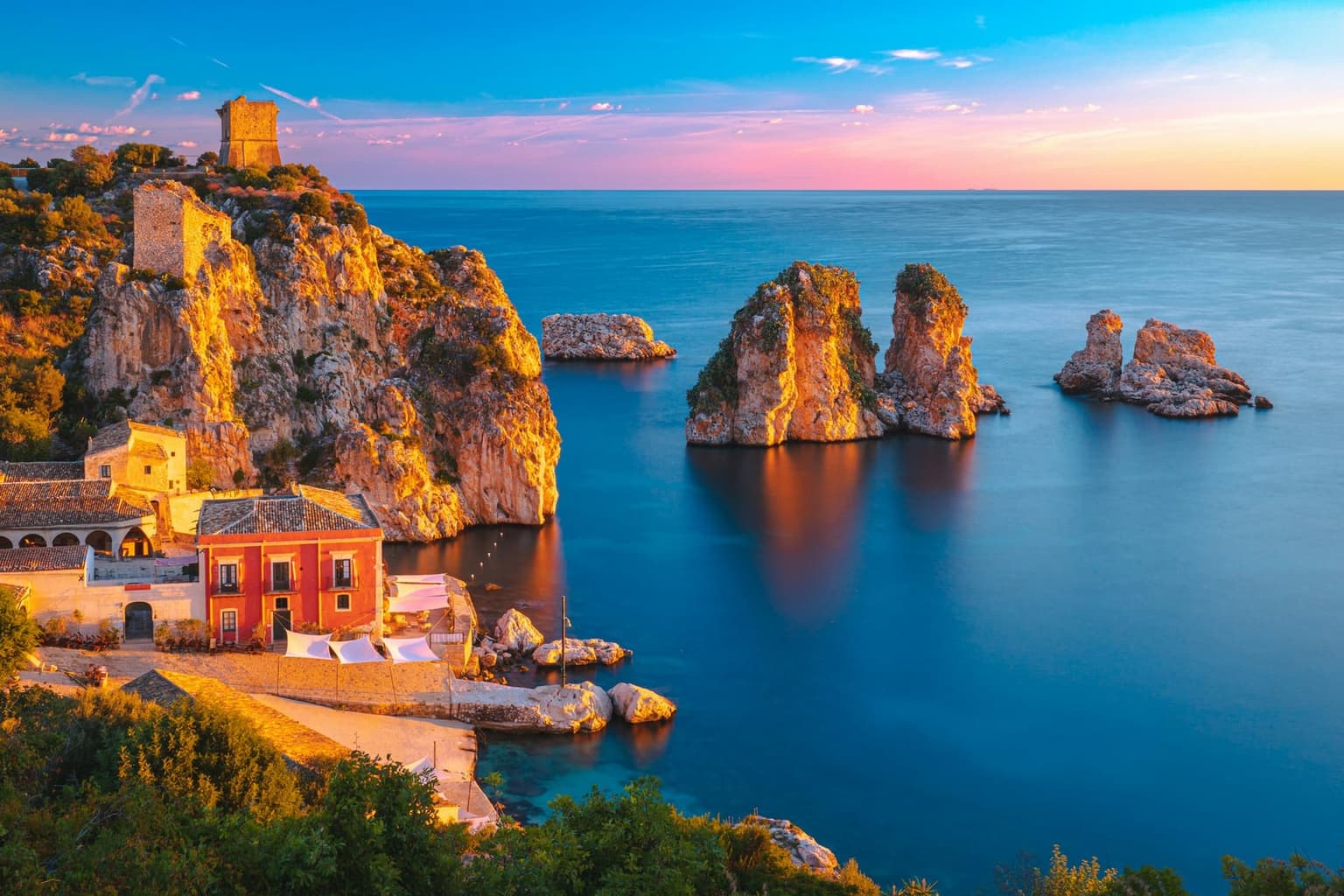 An elevated view of the Scopello cove at golden hour: two tall ochre rock towers offshore, a red-walled tonnara building with a tiled roof and stone watchtower on the cliff above, and a narrow strip of pebble beach between them.