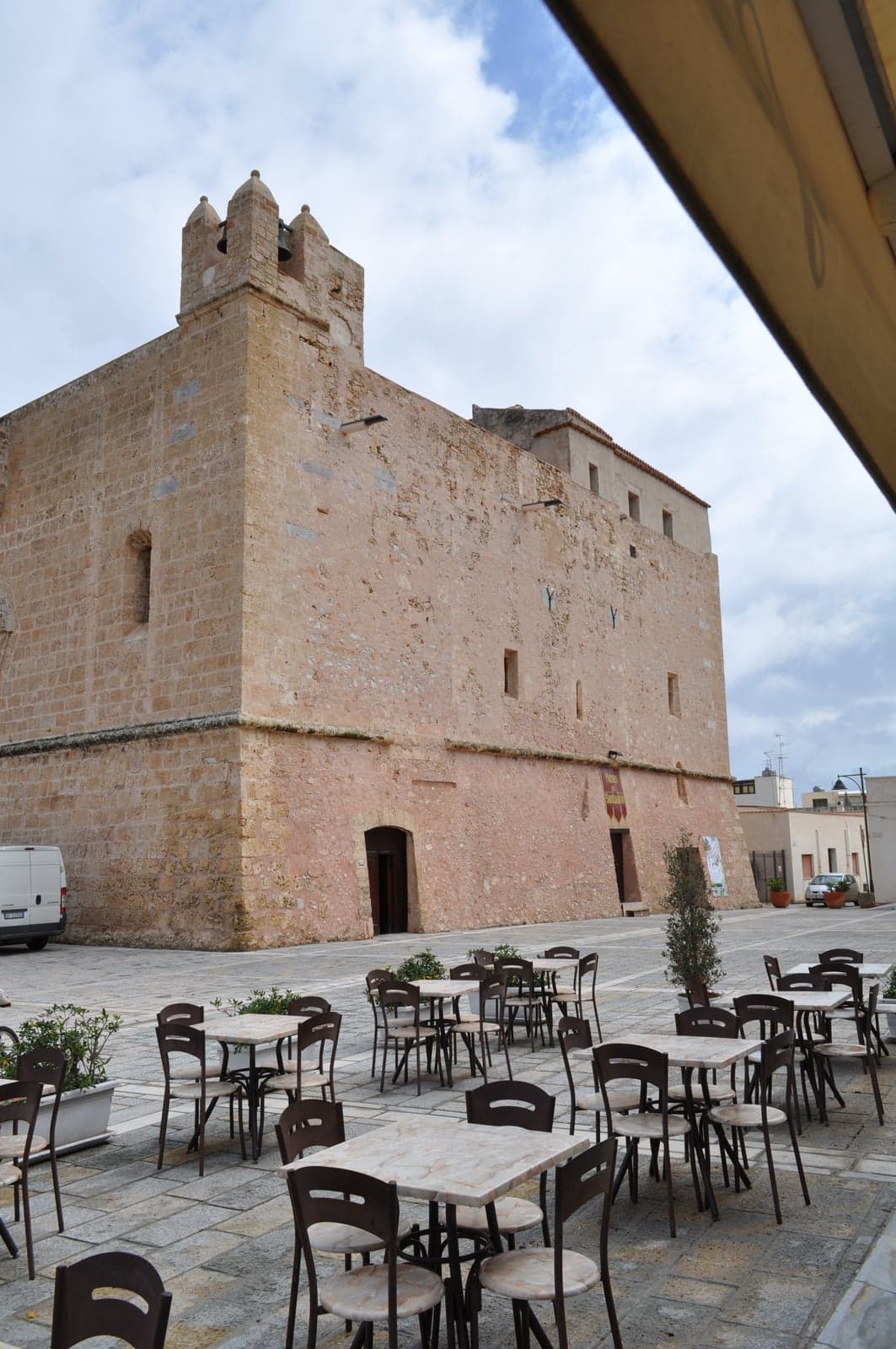 A stone-paved piazza with café tables in front of San Vito's fortress-like sanctuary, its sandstone walls and crenellated bell tower rising above.