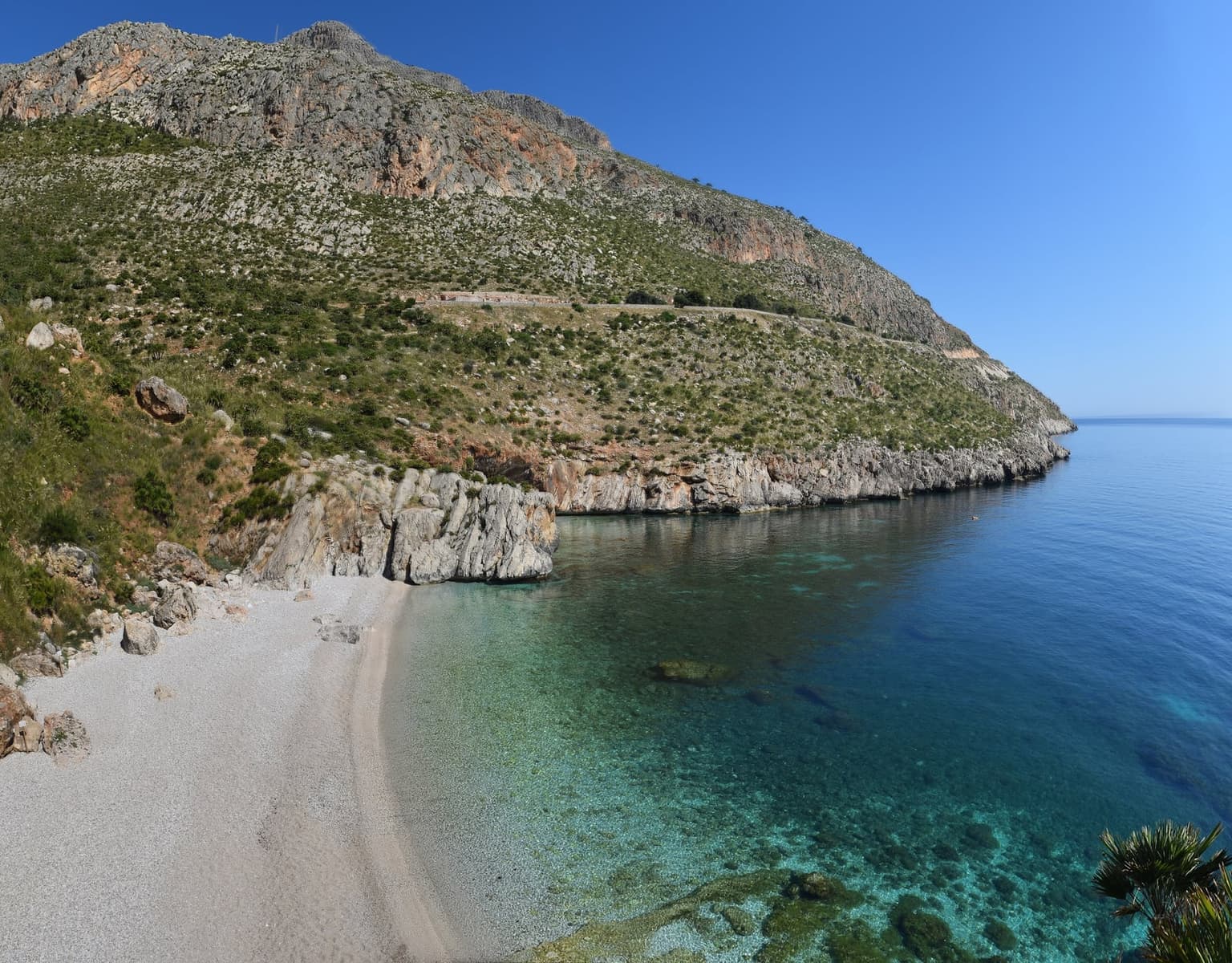 An elevated view down onto a pale pebble cove, the water banding from pale turquoise near shore to deep blue further out, bounded by dry limestone cliffs dropping straight into the sea, with no buildings in sight.
