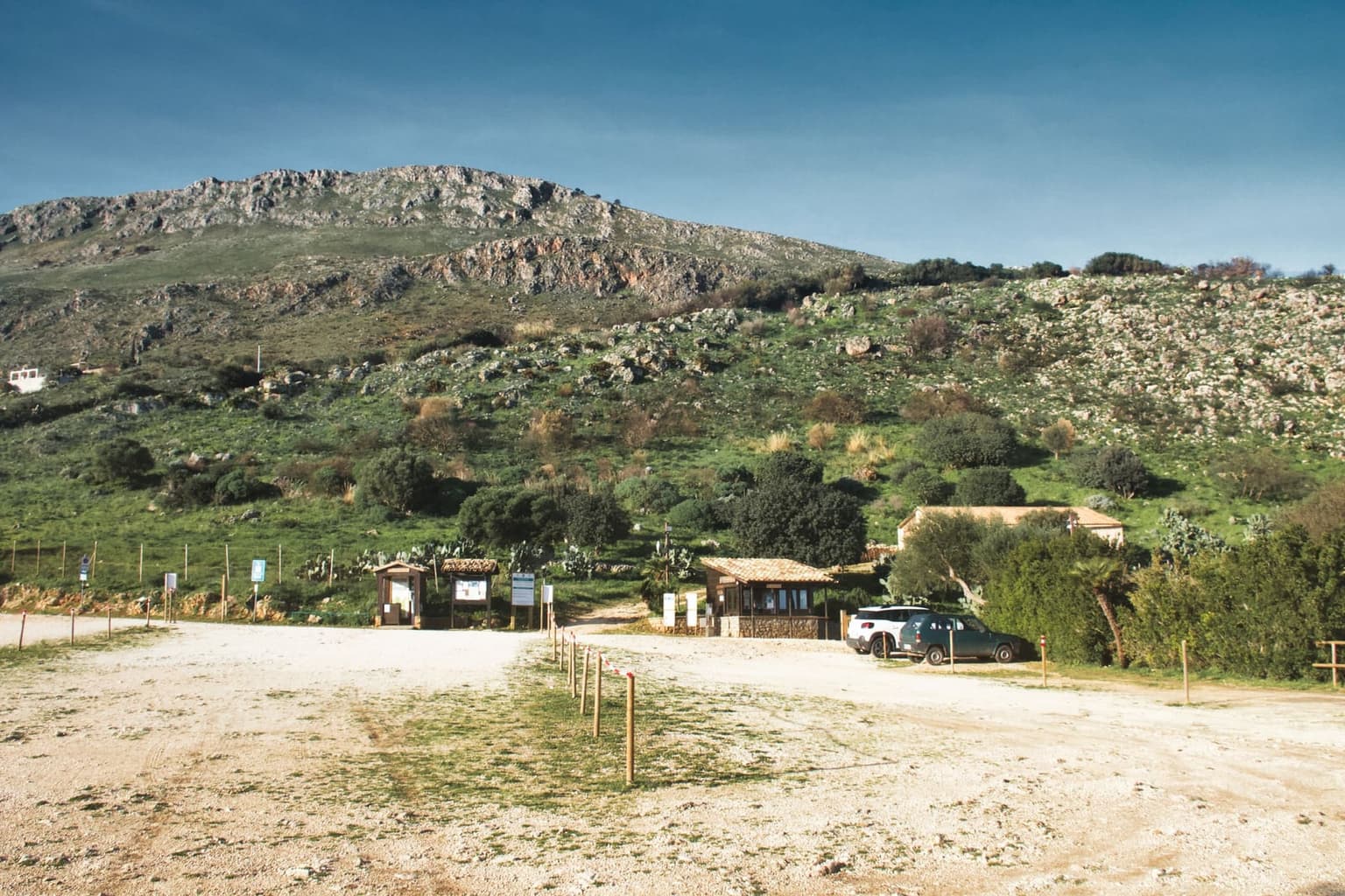 A gravel parking area at the nature reserve's entrance, with a low stone ticket kiosk, information boards, a post-and-tape barrier line, and rocky scrub-covered hills behind.