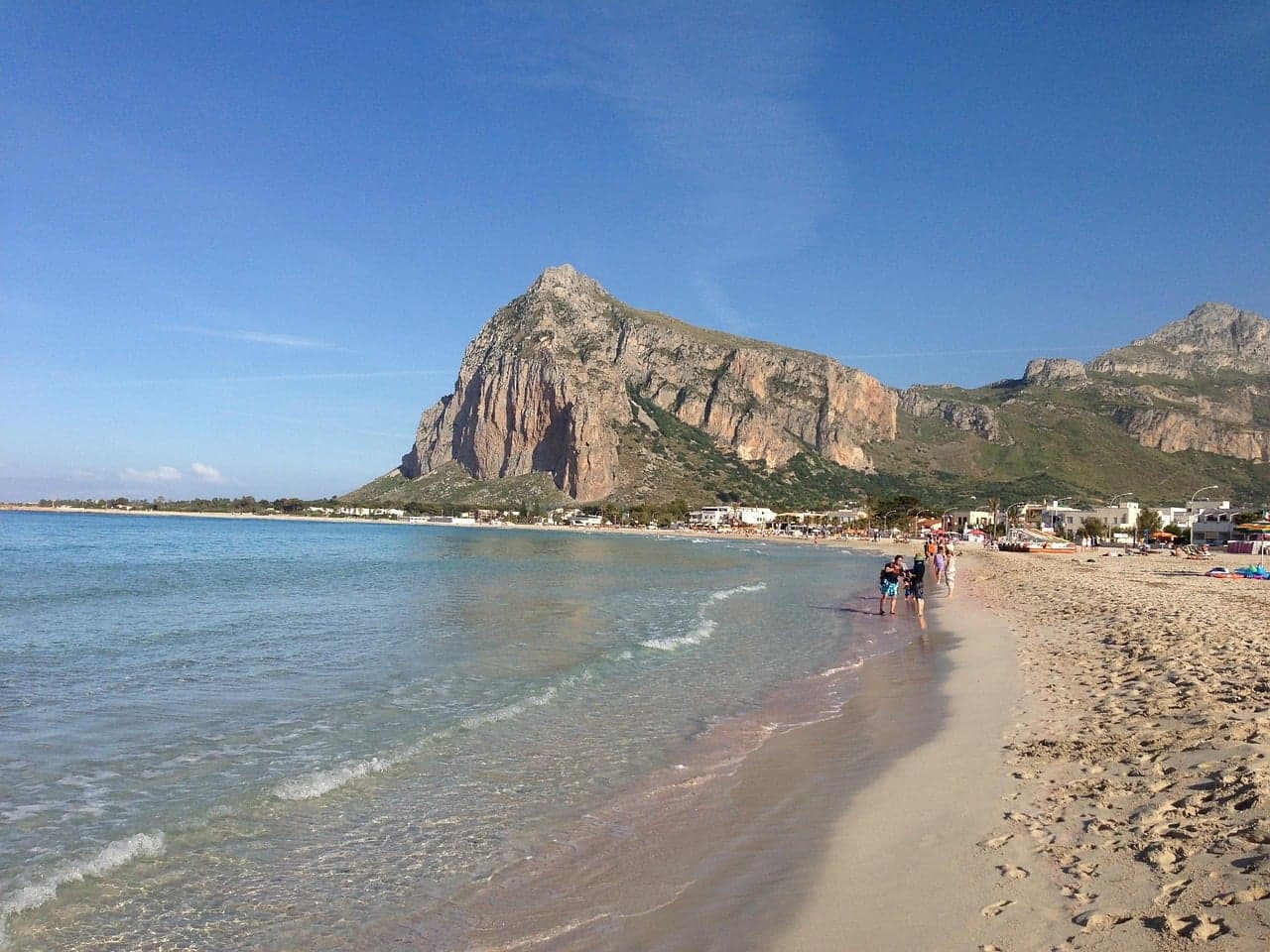 A crescent of pale sand at San Vito Lo Capo with waves lapping the shore, a line of beach umbrellas in the middle distance, and the sheer gray-brown mass of Monte Monaco rising directly behind the town at the water's edge.