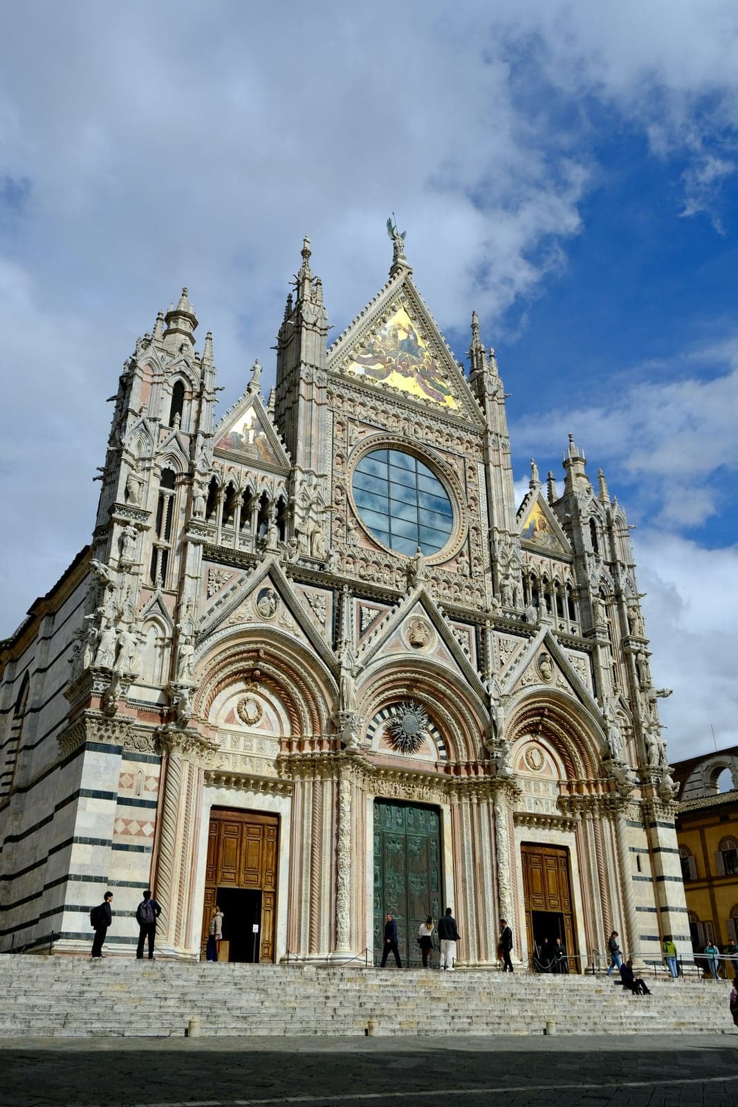 The front of Siena Cathedral in white, pink and dark green marble, with three arched doorways, a large round window, gold mosaics in the pointed gables and black-and-white marble stripes along the side walls.