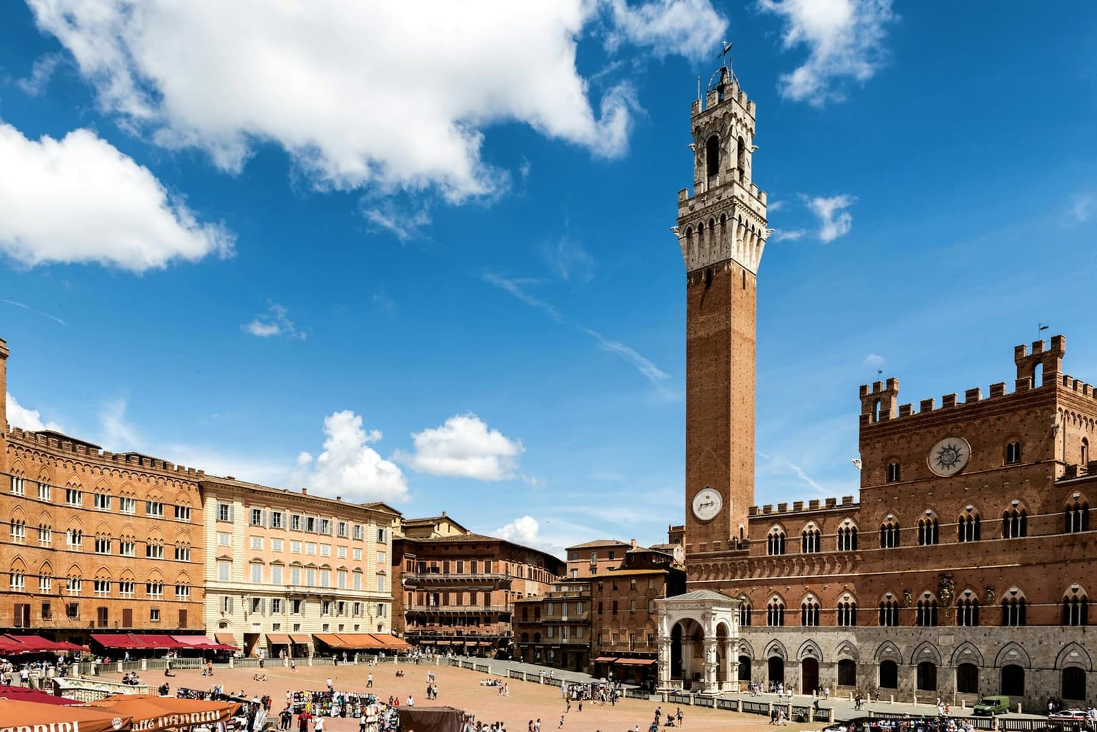Piazza del Campo in Siena seen from above, the fan-shaped brick square sloping toward the crenellated Palazzo Pubblico and the tall brick Torre del Mangia, with a curve of palazzi and red awnings on the other side.