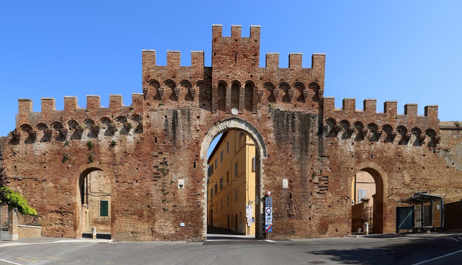 A crenellated brick gate in Siena's walls, with a road leading through the central arch and a column of traffic signs beside it, including a white sign marked zona traffico limitato above a red-ringed circle.