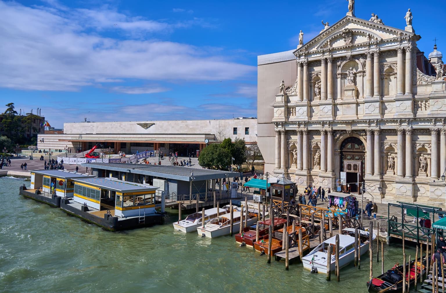 Venezia Santa Lucia station, a long low stone building, faces the Grand Canal across an open plaza, with the yellow-trimmed Ferrovia waterbus stops floating in front, wooden water taxis along the quay and the white facade of the Scalzi church to the right