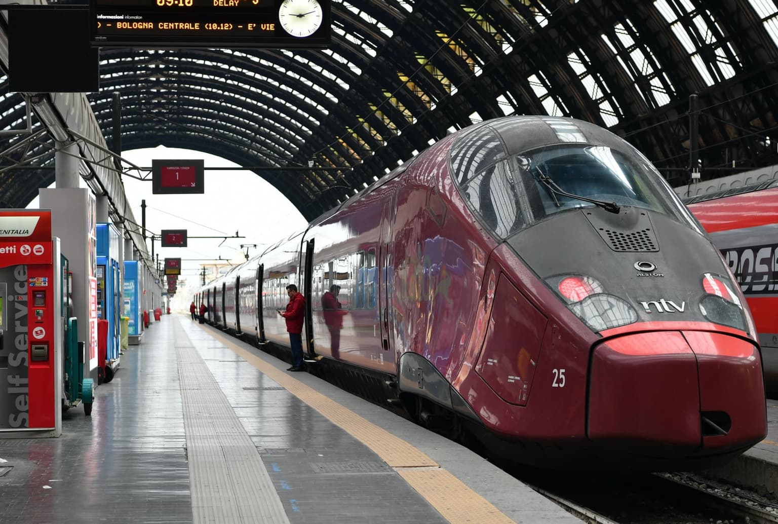 A burgundy Italo high-speed train waits at a platform under the arched iron and glass roof of Milano Centrale, with the side of a red Frecciarossa just visible on the next track