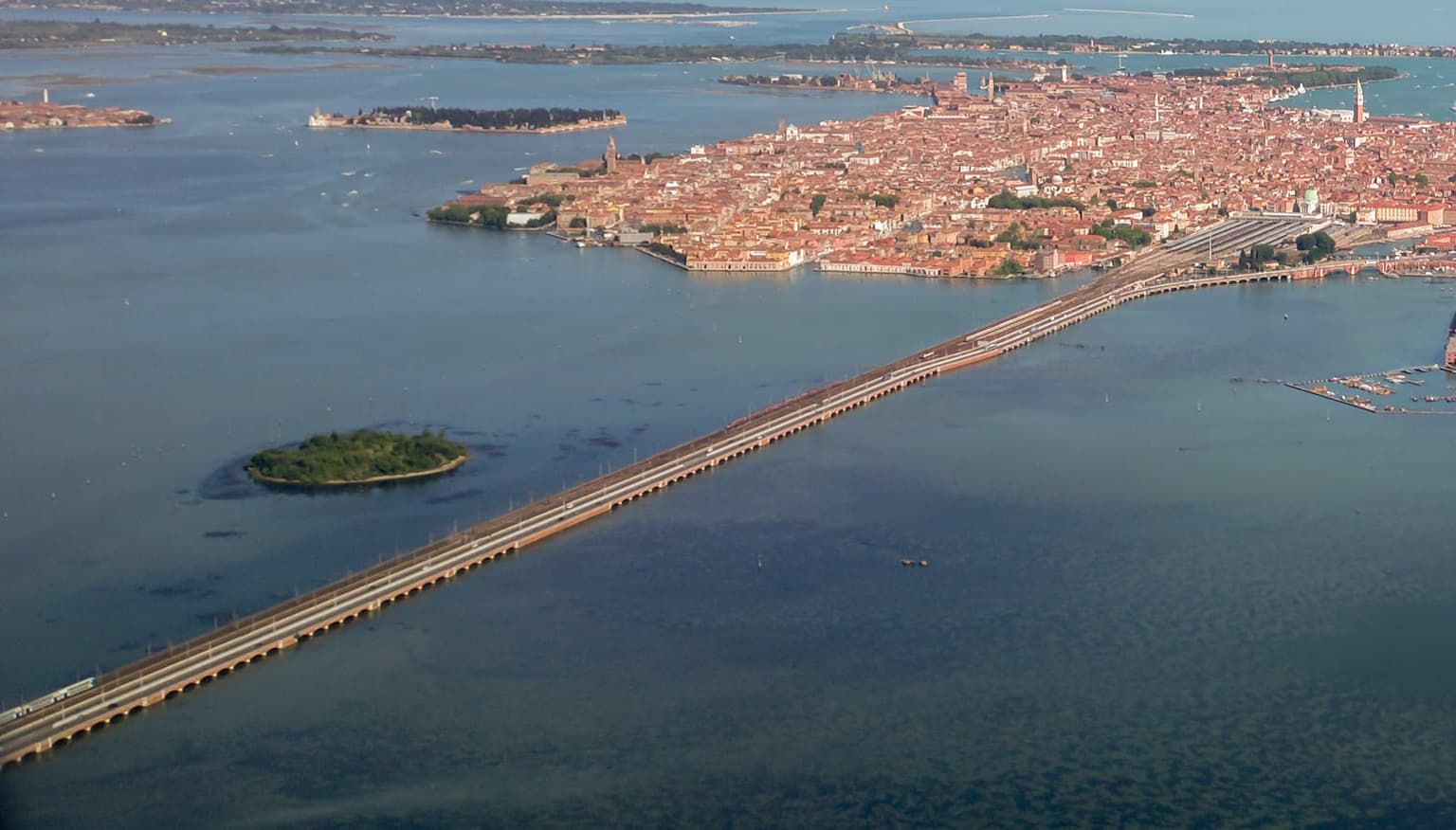 Aerial view of the long rail and road causeway crossing the Venetian lagoon to Venice, with a train on the bridge in the foreground, the station's track yard where it reaches the city, and the red-roofed historic center beyond