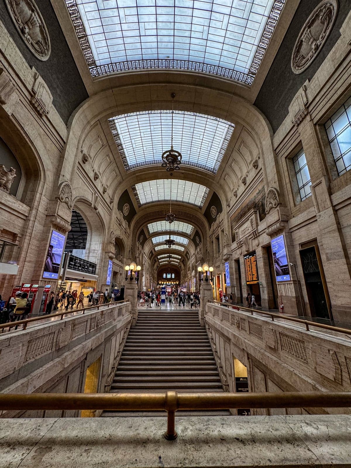 Inside Milano Centrale, a wide stone staircase leads up to a long vaulted gallery with glass skylights and hanging lanterns, with travelers crossing the concourse, red ticket machines on the left and a departure board on the right